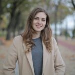 A young woman in a beige coat smiles warmly while standing outdoors during fall.