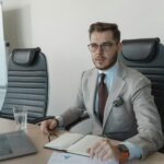 Businessman wearing suit and glasses working at desk with laptop, notebook in office.